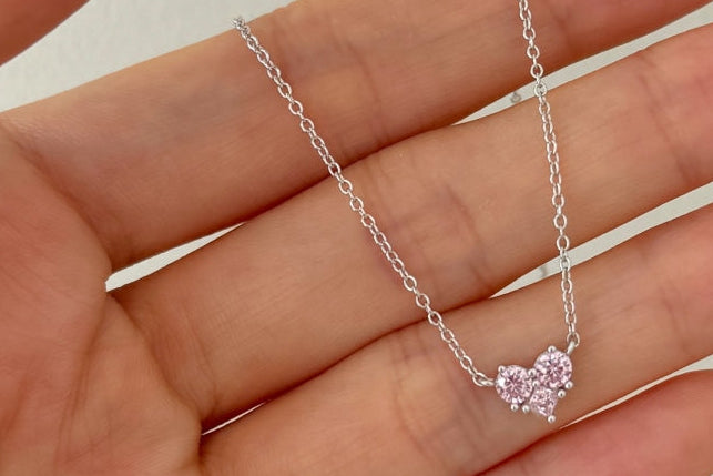 Hand holding a silver necklace with a heart-shaped pink pendant against a plain background